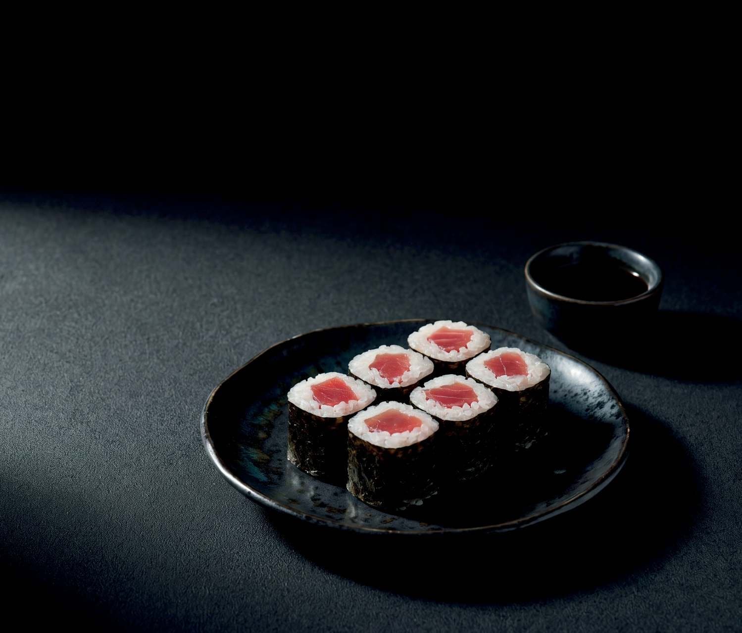 Six maki au thon sur une assiette noire, un petit bol de sauce soja derrière, dans la pénombre